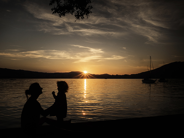 Foto: Wilhelm Watschka Fotografie. Mutter und Kind vor einem See als Silhouette. Hinter den Bergen geht gerade die Sonne unter. Aufgenommen am Attersee - Oberösterreich