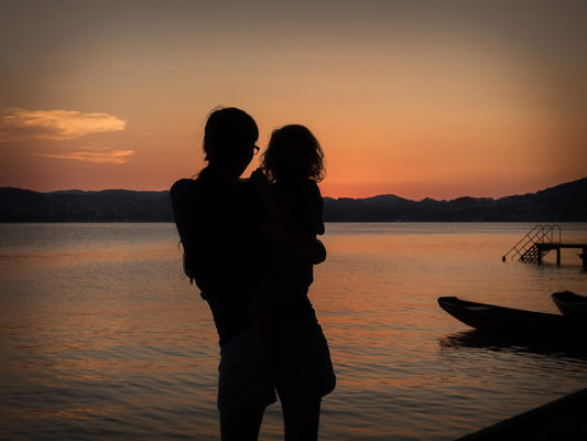 Eine Frau hält ihre Tochter im Arm und sieht sich den romantischen Sonnenuntergang am See an. Foto von Wilhelm Watschka