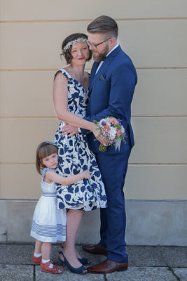 Ein Paar steht nach der Hochzeit vor einer Wand und posiert nach den Anweisungen des Fotografen. Plötzlich läuft die Tochter ins Bild und hält sich bei der Braut fest. Foto von Wilhelm Watschka