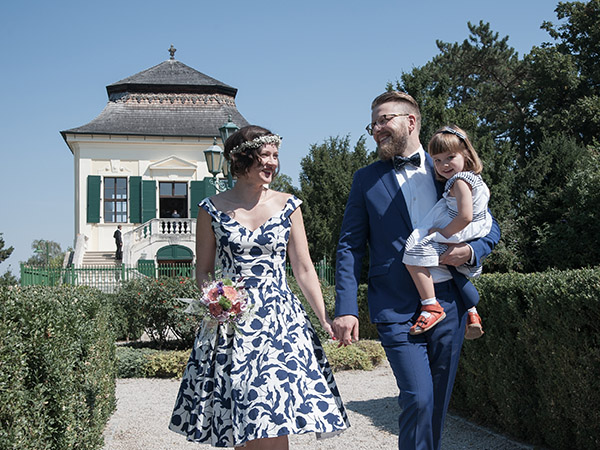 Foto: Wilhelm Watschka Fotografie. Nach der Hochzeit spaziert ein Paar mit ihrer Tochter im Arm den Park entlang