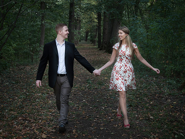Foto: Wilhelm Watschka Fotografie. Ein verliebtes Paar spaziert Händchenhaltend durch einen Laubwald und strahlt sich dabei an.