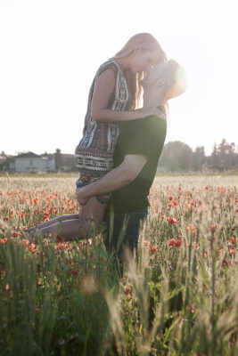 Ein Paar steht im Mohnblumenfeld im Gegenlicht. Er hebt seine Freundin vorsichtig hoch und sie küssen sich dabei. Foto wurde von Wilhelm Watschka gemacht