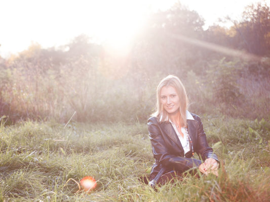 Foto: Wilhelm Watschka Fotografie I Eine Frau sitzt in einer Wiese und hinter ihr geht die Sonne unter. Im Bild sind Blendflecken zu sehen. Traumhaft schön!